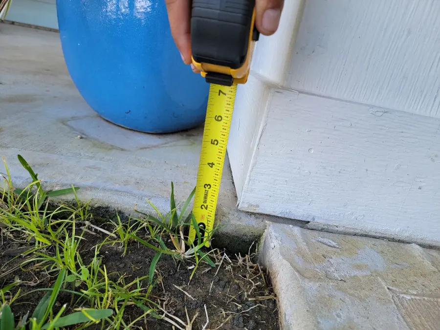 A tape measure held upright at the edge of a concrete slab and siding, measuring height above the soil of a planting bed